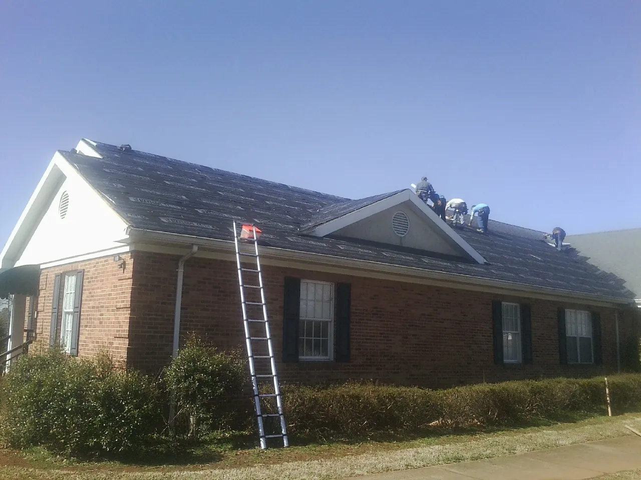 Crew tearing shingles off a brick ranch roof with a ladder set — Metal Roof Repair work by our crew in Issue